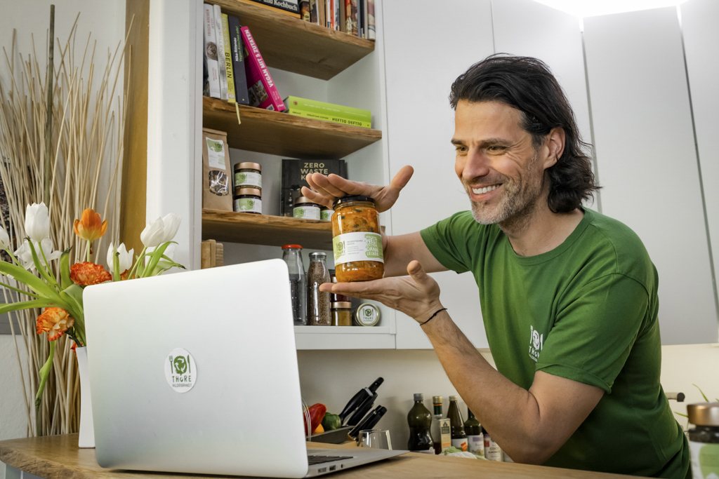 Mann im grünen Shirt zeigt in seiner Küche ein Glas Kimchi, davor ein Laptop