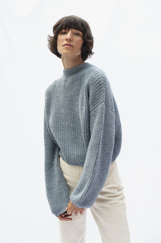 Frau im blaugrauen Strickpullover und heller Hose vor weißem Hintergrund