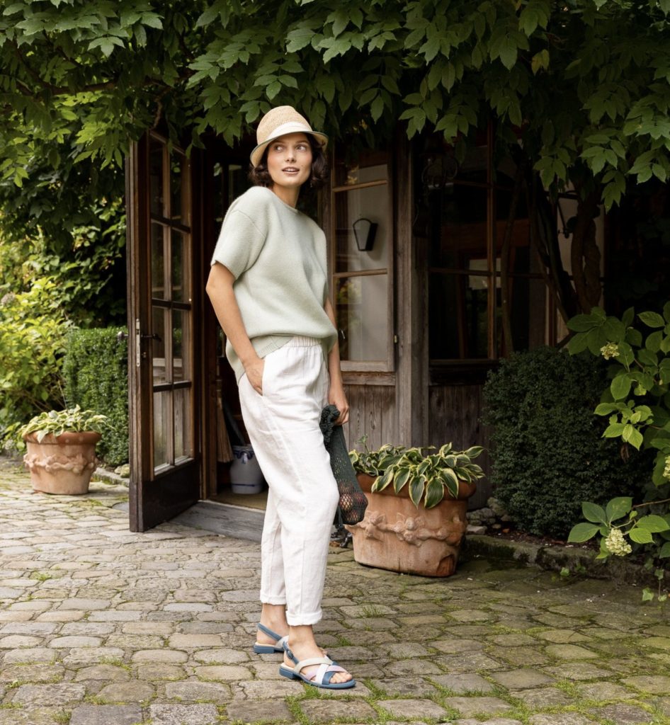 Frau mit Fischerhut und weißer Hose vor einem Gartenhaus