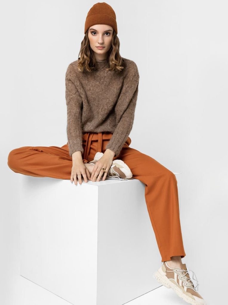 Frau in braunem Pullover und orangefarbener Hose auf einem weißen Würfel