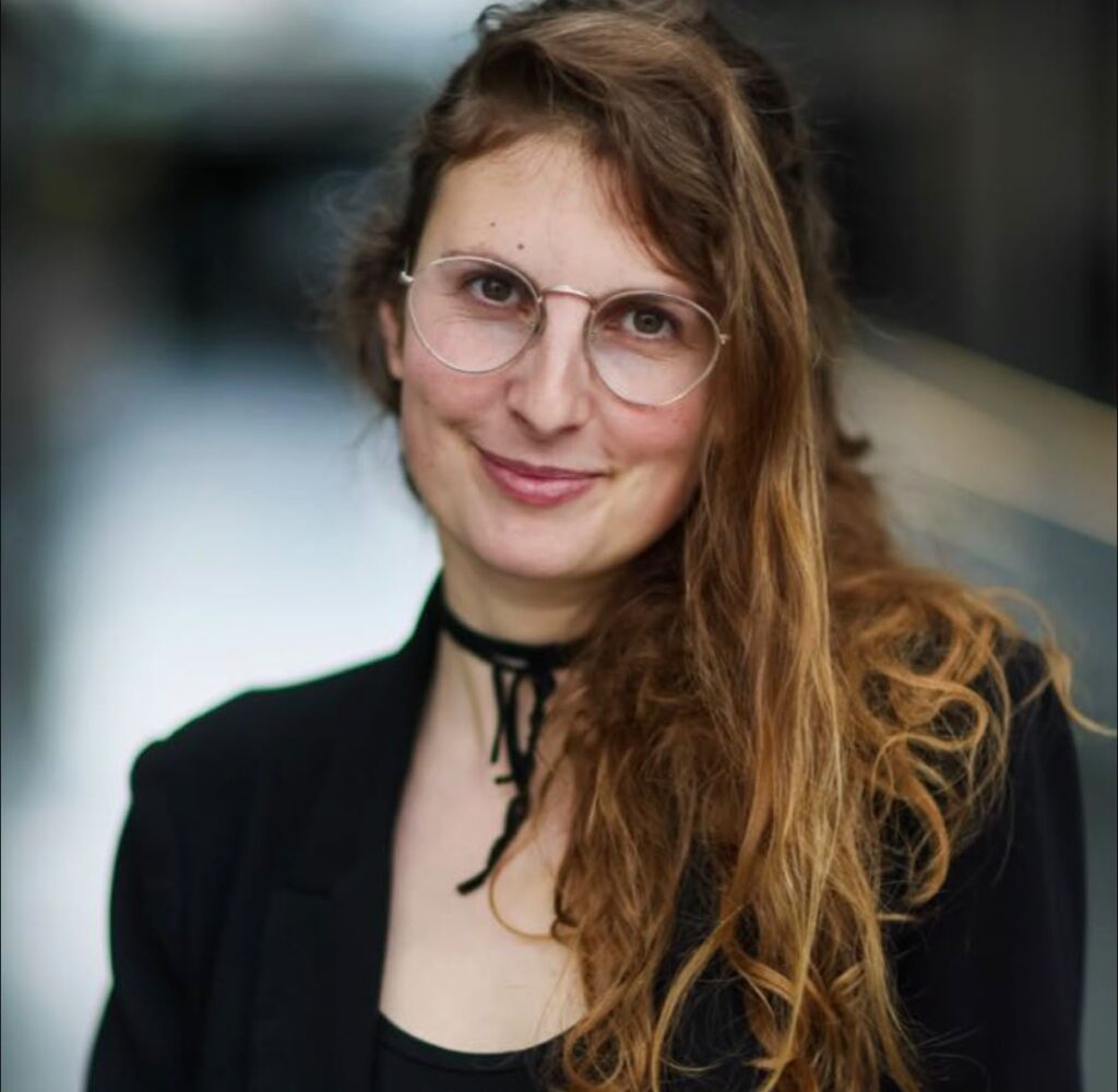 Anna mit langen, gewellten Haaren und runder Brille im schwarzen Blazer