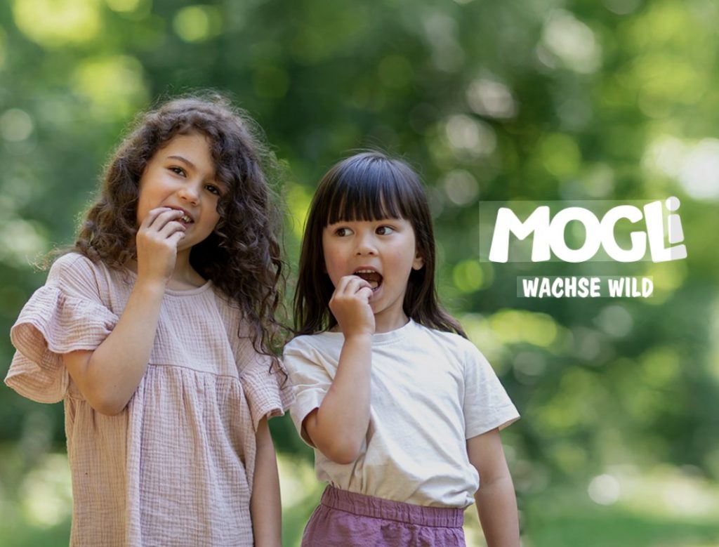 Zwei Mädchen essen Snacks im Grünen, daneben das Logo „Mogli – Wachse wild“