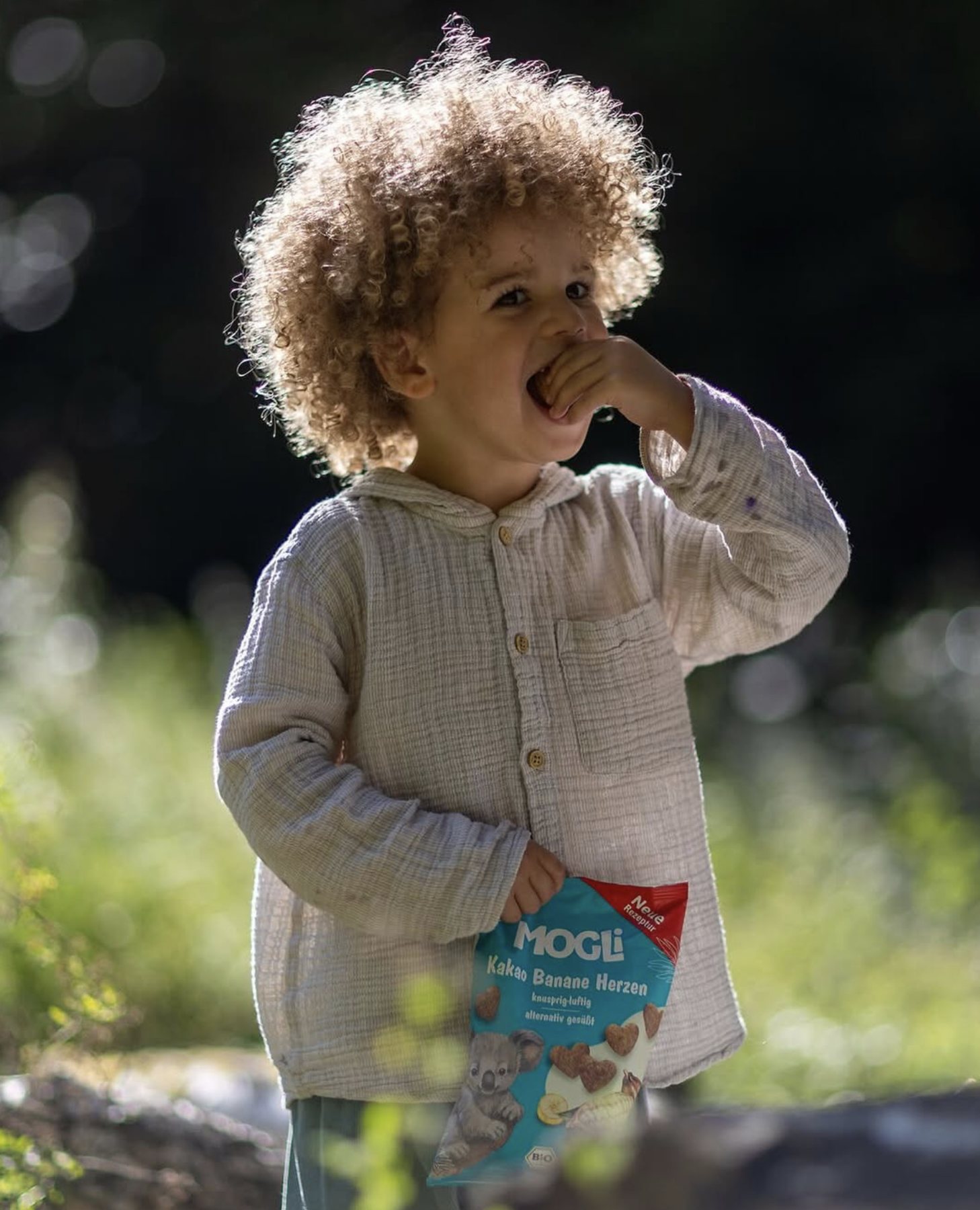 Kleinkind mit Locken isst im Grünen einen Snack von Mogli