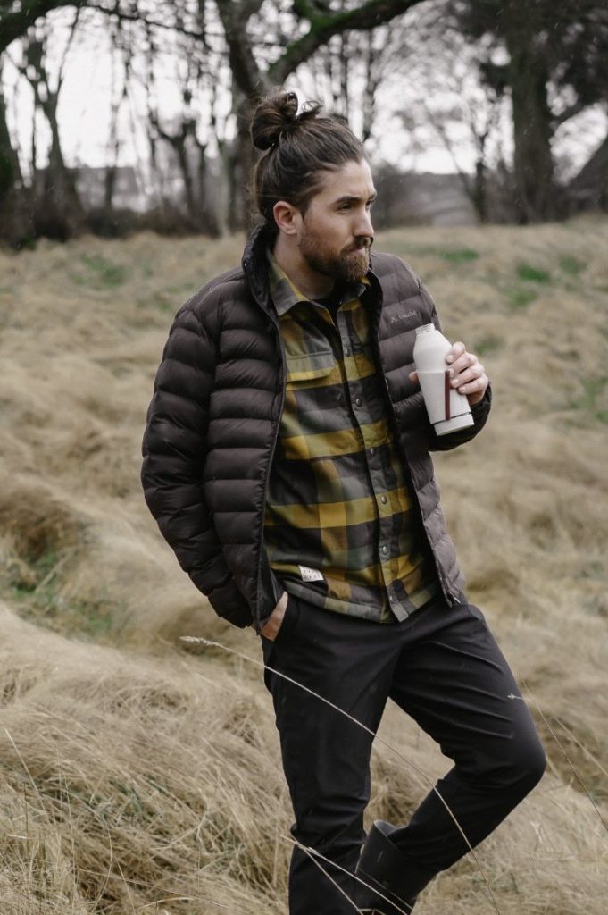 Mann in Steppjacke und kariertem Hemd mit Trinkflasche im hohen Gras